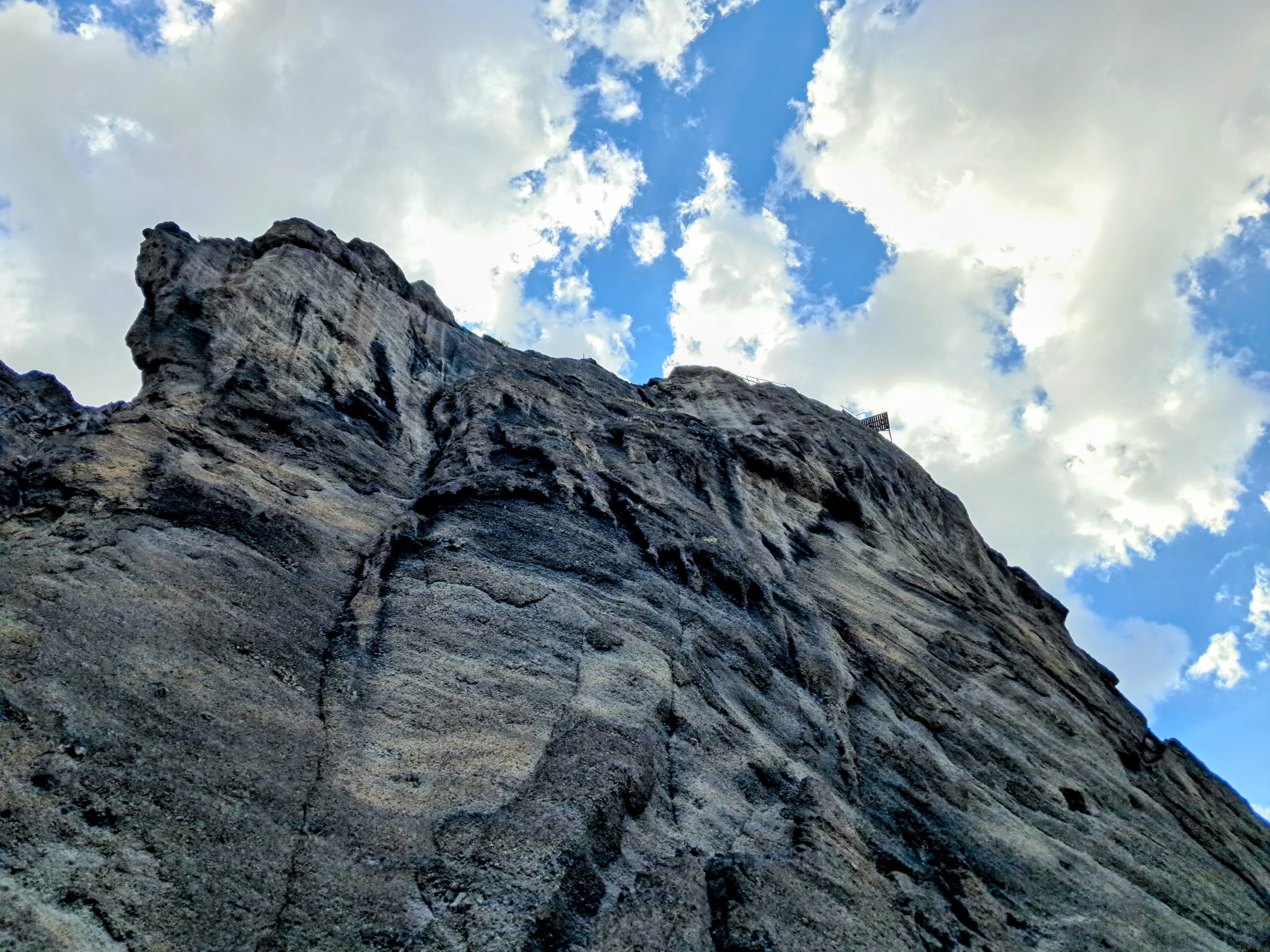 A low-angle view of a massive rugged dark gray rock cliff against a blue sky with scattered white clouds. The steep cliff face displays rough texture, deep crevices, and natural striations. Near the upper ridge a ramp, used for bungee jumping or paragliding, is faintly visible along the skyline.