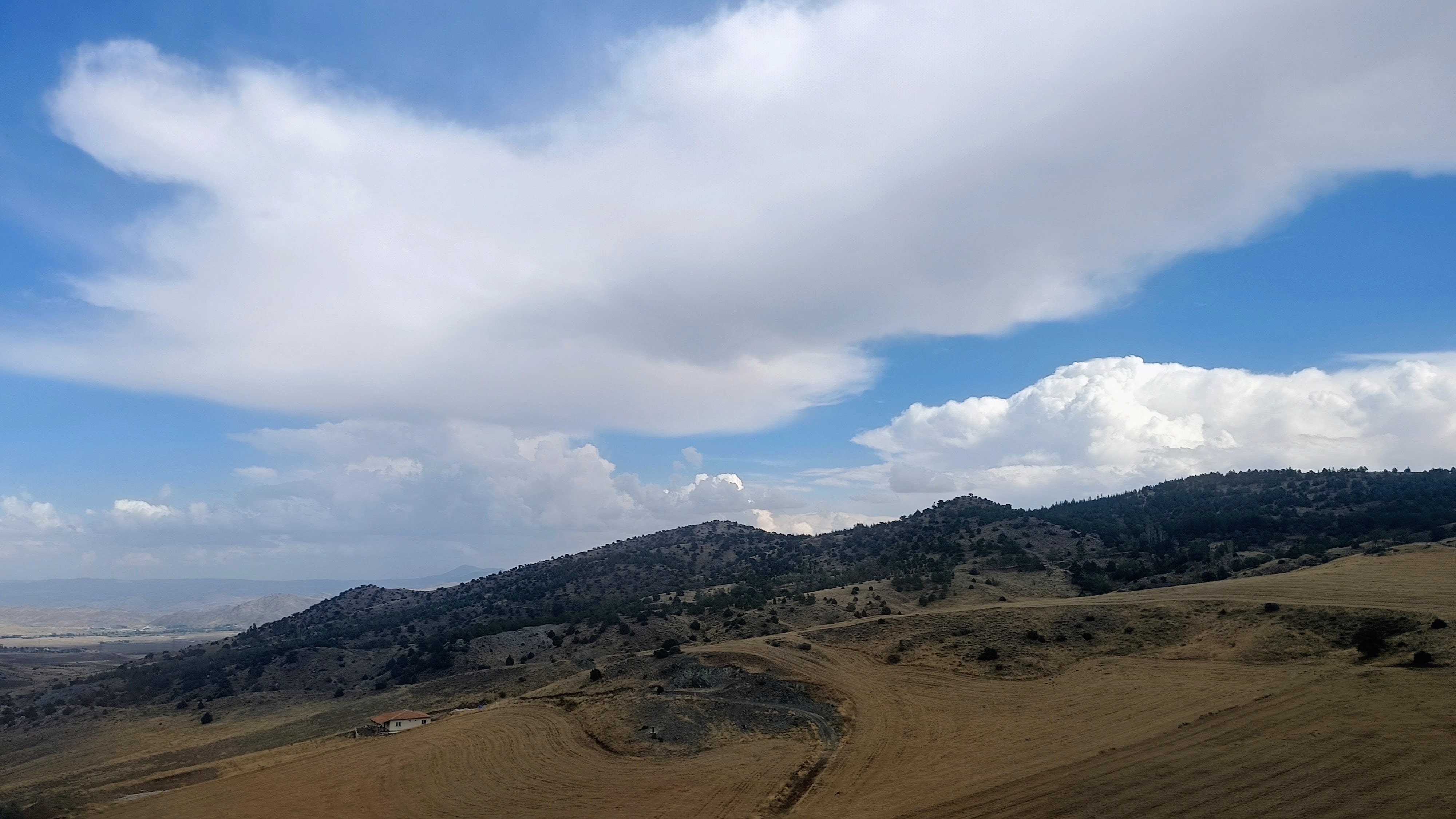 A wide landscape with rolling golden hills in the foreground leading up to dark tree filled ridges in the distance under a blue sky with wide white clouds. In the lower left among the fields sits a single small building with a red-tiled roof. Large cloud formations cover the upper half of the frame, casting shadows across parts of the open terrain.