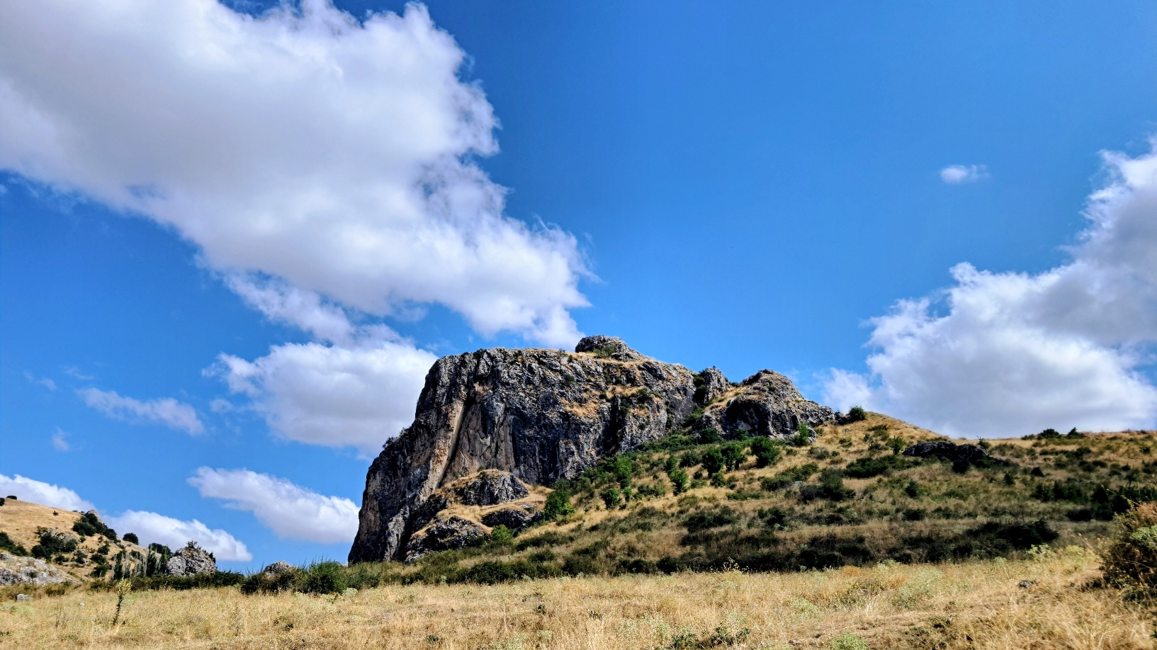 A rugged rocky cliff rises abruptly from a dry grass meadow with a few scattered green bushes. The sheer rock face features craggy weathered gray stone that contrasts with the surroundings arid hillside, all under the blue sky filled with fluffy white clouds and sunlight.