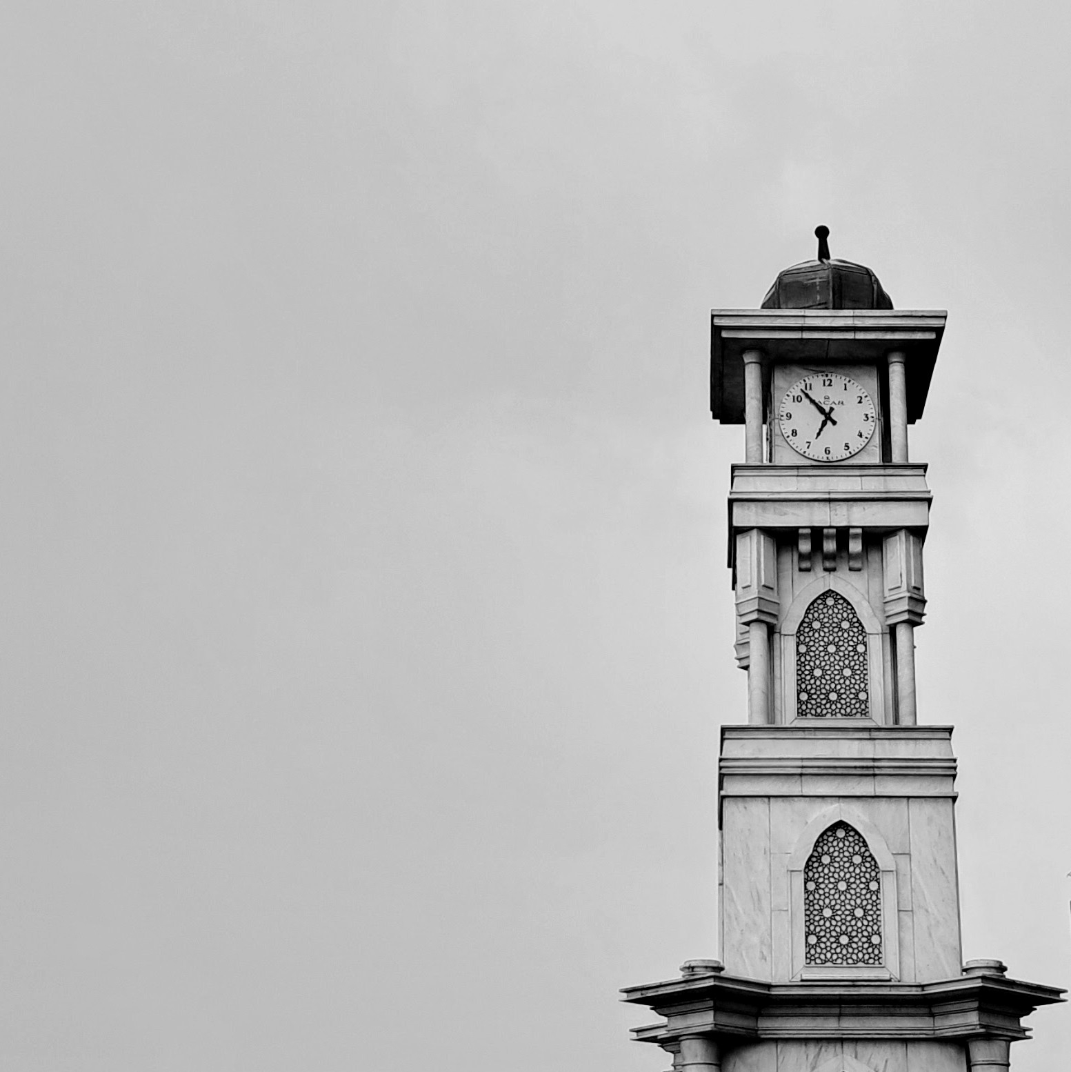 A multi-level ornate stone clock tower against an overcast gray sky. The upper section features a round analog clock face displaying standard numerals showing 6:52. Below the clock, the tower has arched vertical niches inlaid with intricate geometric latticework, all covered under a dark domed roof and finial.