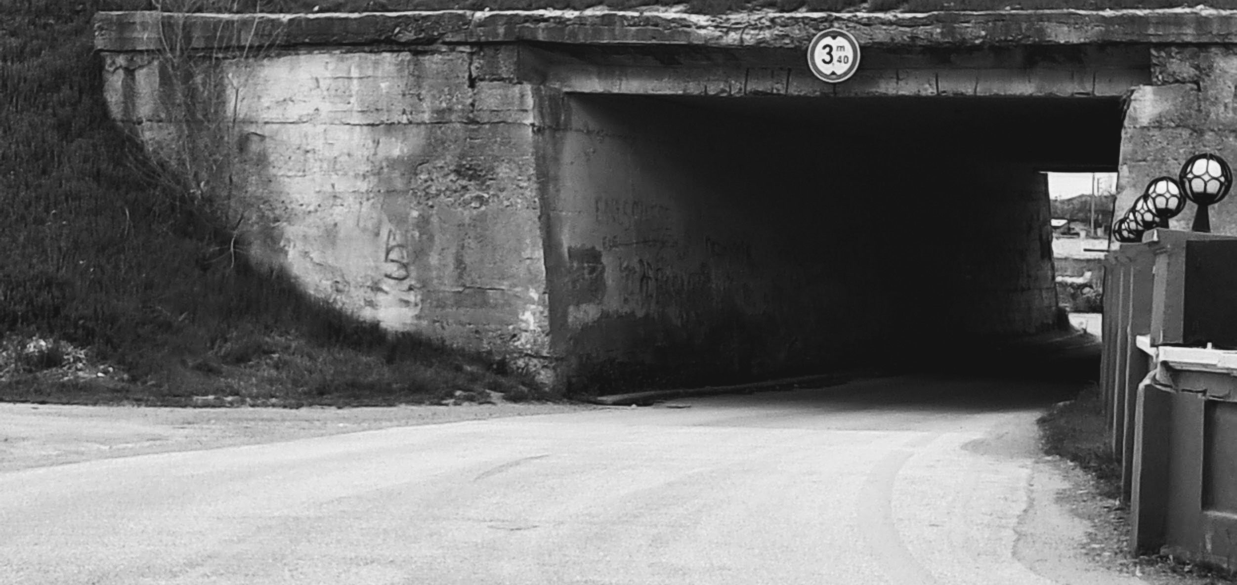 A paved road leading into a dark concrete underpass beneath an overpass. A circular height clearance sign mounted above the center of the entrance reads "3 m 40" with vertical arrows. Weathered concrete walls with faint illegible graffiti frame the tunnel opening, bordered by a sloping embankment on the left and a low roadside barrier with several decorative spherical lamps on the right. The road curves through the shadowy interior toward a bright exit in the distance.