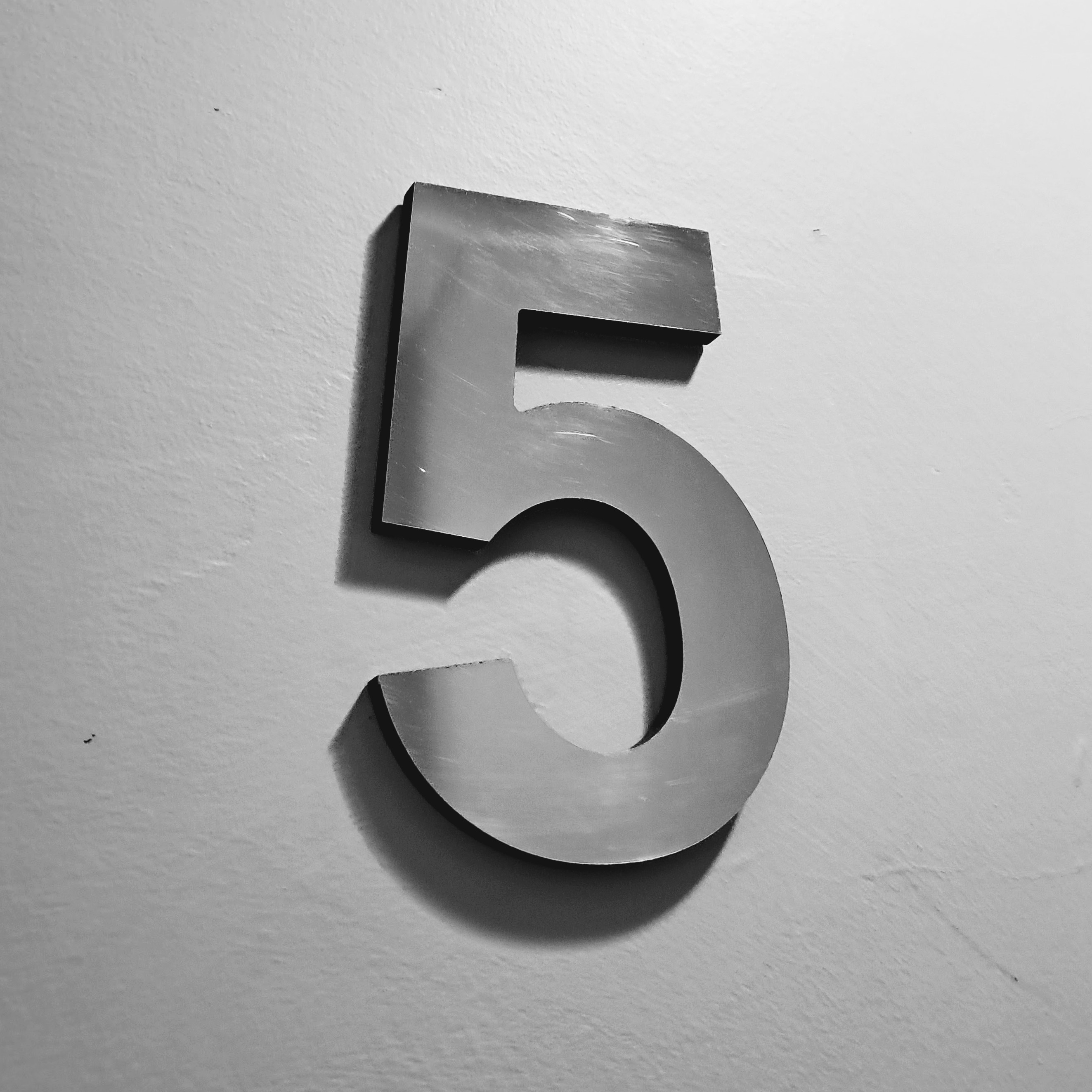A three-dimensional number 5 made of brushed metal mounted on a white textured wall. Strong lighting from the upper right casts a shadow beneath and to the left of the numeral.