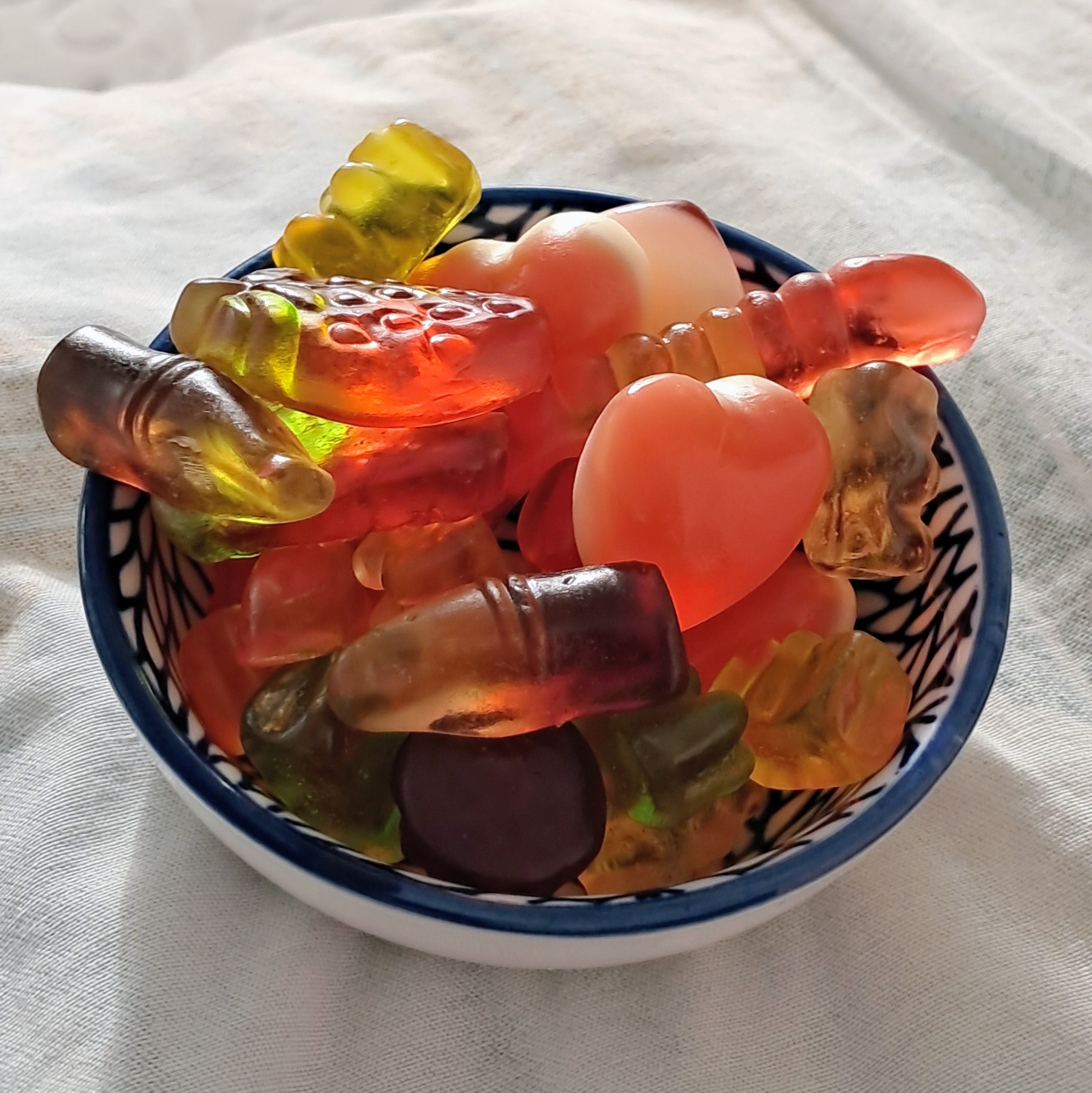 A small ceramic bowl with a blue rim and patterned design is filled with an assortment of gummy candies on a white fabric surface. The variety includes translucent cola bottles, gummy bears, a worm, a textured fruit shape resembling a strawberry, and opaque pink heart-shaped candies. Soft natural light illuminates the mix, highlighting the translucent textures and vibrant shades of red, yellow, pink, and brown.