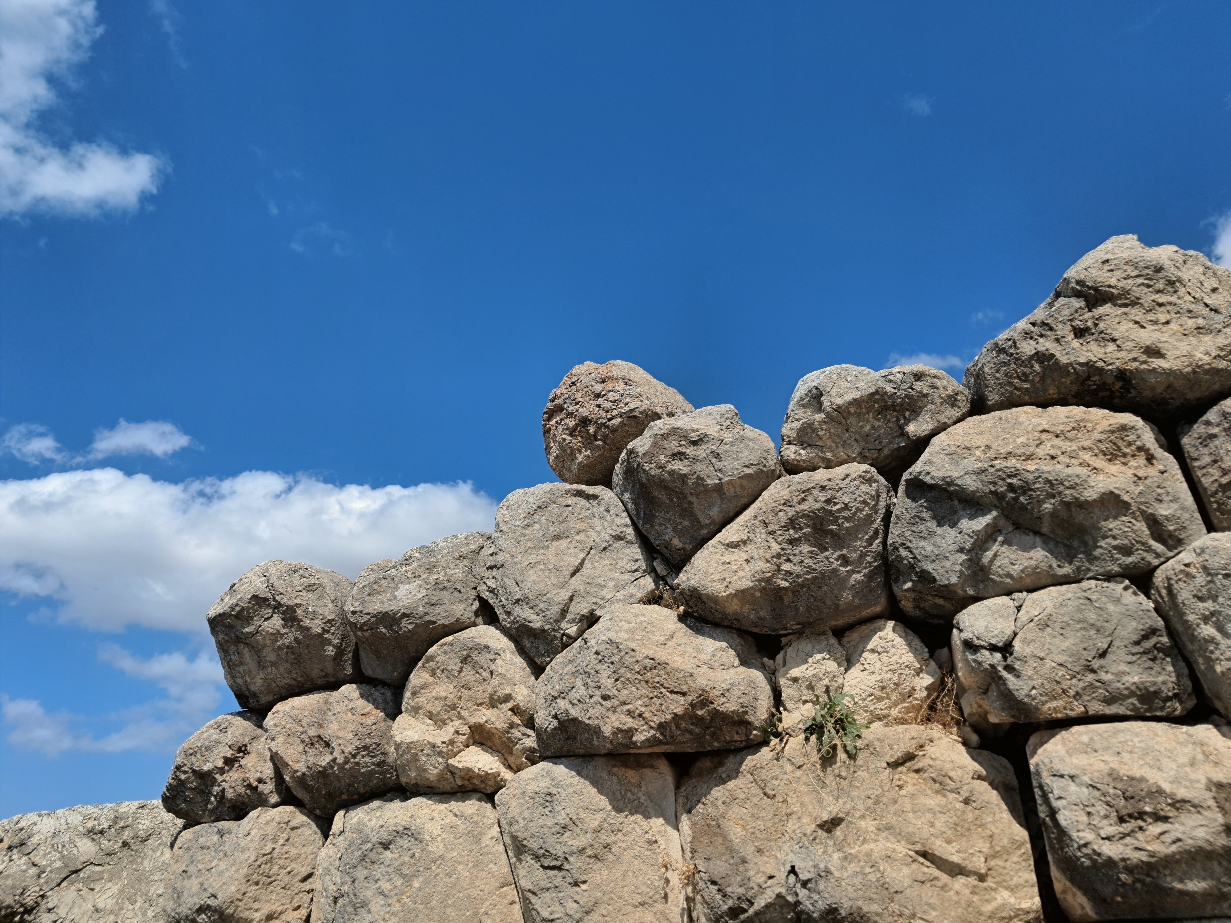 An ancient dry-stone wall made of large interlocking gray boulders stacked together without visible mortar. A tiny green plant sprouts from a crevice between the stones. Above the uneven stone structure, a blue sky with scattered white clouds on the left is visible.