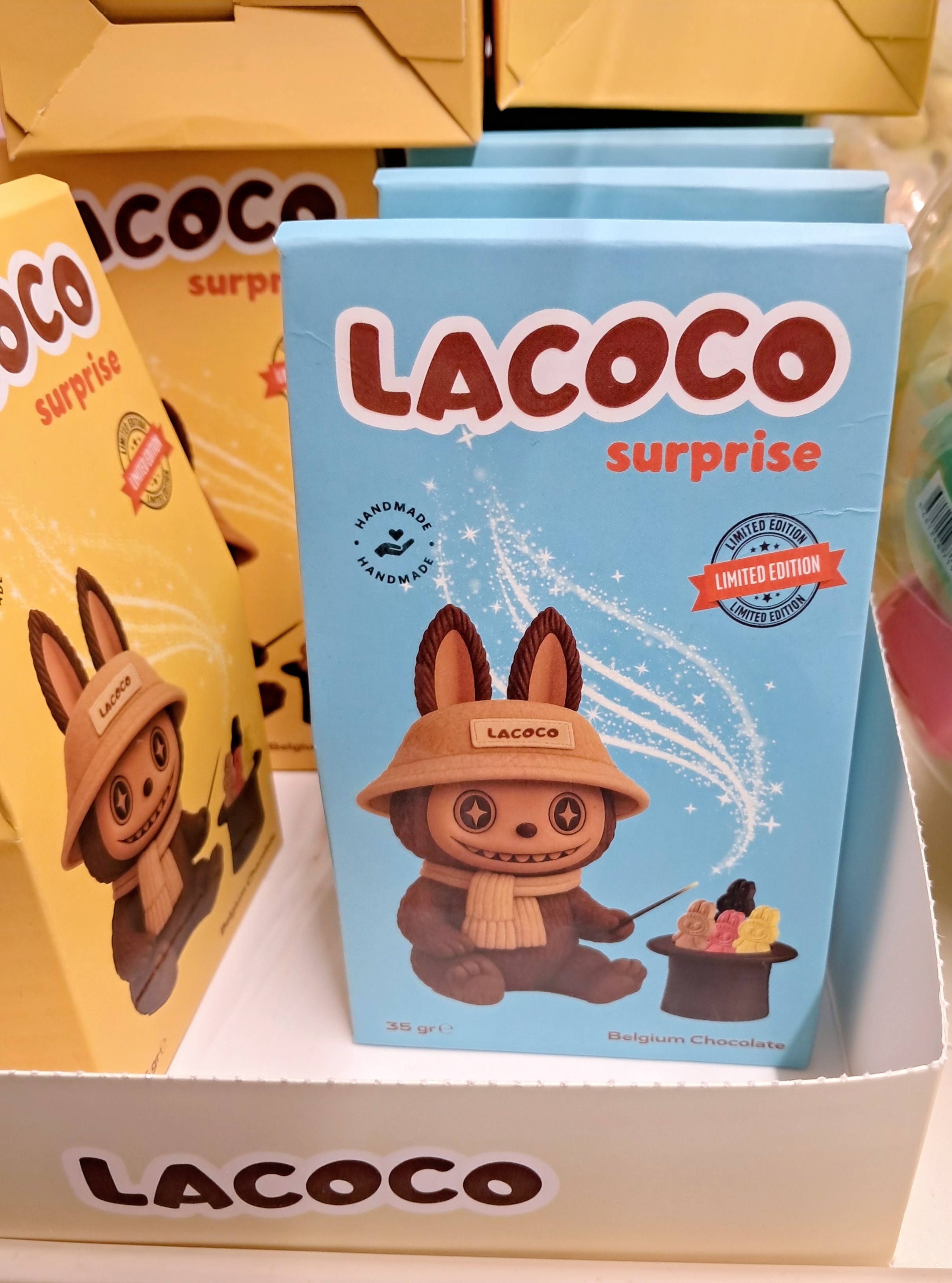 A light blue "LACOCO surprise" chocolate box features an illustration of a brown rabbit-eared character resembling a Labubu™ creature wearing a beige bucket hat with the text "LACOCO" and a matching knit scarf. The character smiles broadly with pointed teeth and holds a small wand over a black top hat, from which colorful gummy-like figures emerge amid magic light. The front displays circular badges reading "HANDMADE" and an orange ribbon stamp marking it as "LIMITED EDITION." Small text at the bottom reads "35 gr" alongside "Belgium Chocolate", sitting in a cardboard display tray with additional yellow and blue boxes visible in the background.